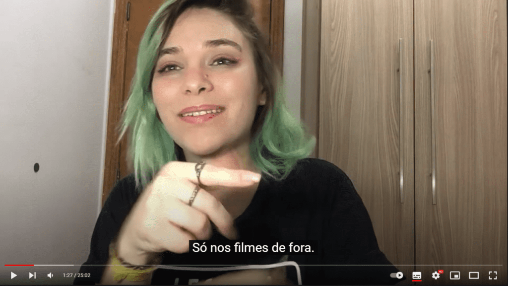 Imagem retirada do vídeo “COMO É A ACESSIBILIDADE PARA SURDOS NA PANDEMIA?” que apresenta Flávia Lima, tradutora e intérprete de Libras. Mulher branca com cabelos descoloridos e verde. Usa a camisa da campanha “Legenda para quem não ouve, mas se emociona”. No trecho da cena, a legenda: "Só nos filmes de fora."