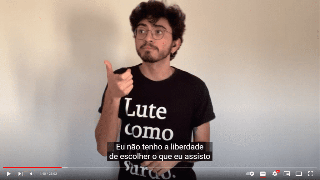 Imagem retirada do vídeo “COMO É A ACESSIBILIDADE PARA SURDOS NA PANDEMIA?” que apresenta Gabriel Isaac, YouTuber e designer gráfico. Homem branco, de cabelos lisos, pretos e desgrenhados. Usa óculos de grau, bigode, barba e uma camiseta preta contendo a seguinte frase: "Lute como um surdo." No trecho da cena, a legenda: "Eu não tenho liberdade de escolher o que assisto..."