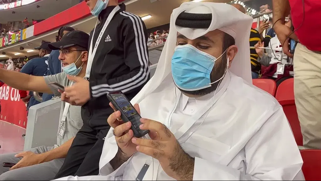 Homem de turbante branco e máscara segura o smartfone utilizando o aplicativo FIFA Interpreting disponibilizado pela FIFA realizando testes de utilização.