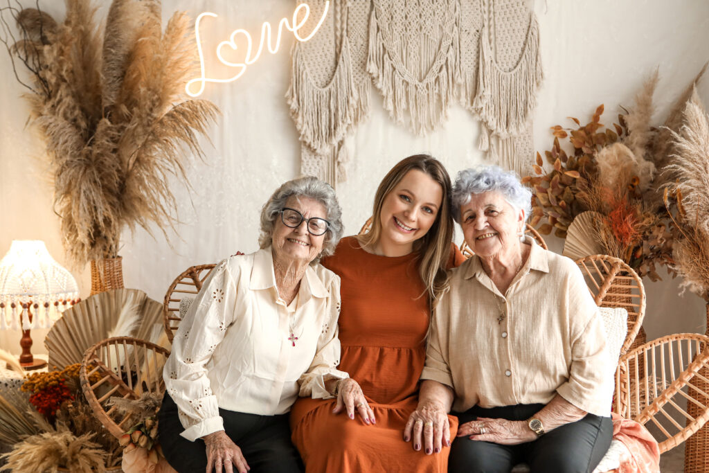 Tita, Kaine e Vó sentadas uma ao lado da outra, sorrindo. Tita e Vó vestem branco, enquanto Kaine veste vermelho.